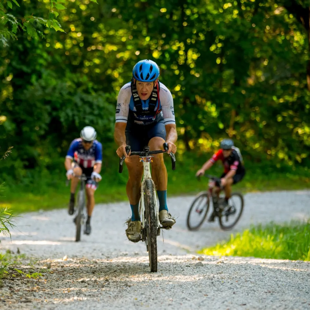 Three cyclists riding on a gravel path through a sunlit forest, with the lead cyclist focused and wearing a blue helmet.