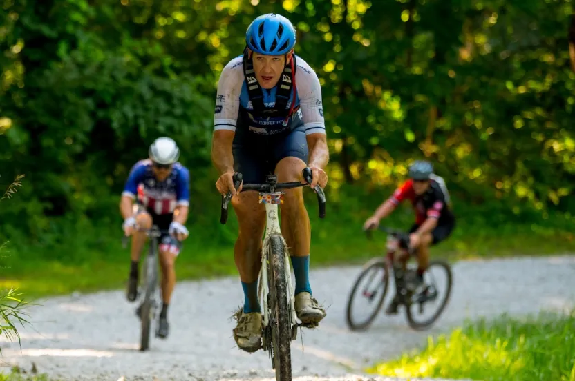 Three cyclists riding on a gravel path through a sunlit forest, with the lead cyclist focused and wearing a blue helmet.