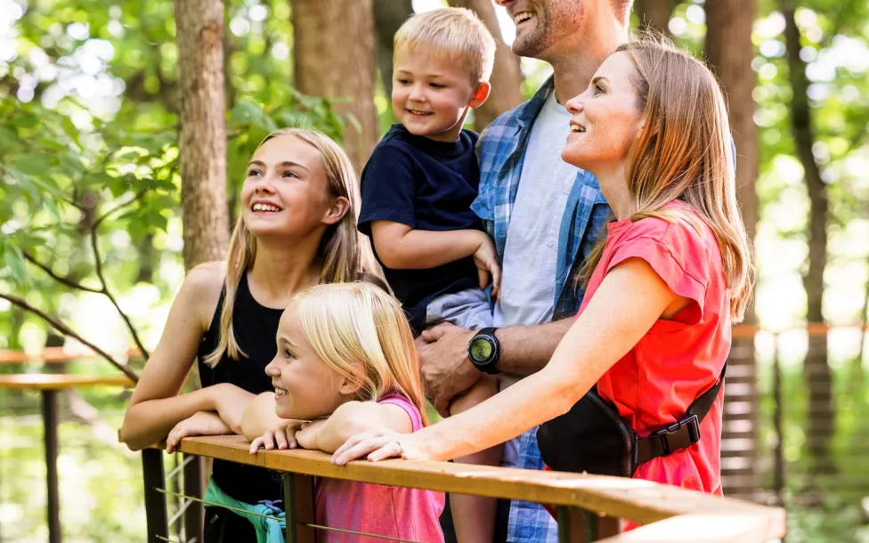 Family of five with two adults and three children standing on a wooden platform surrounded by green trees in a park.